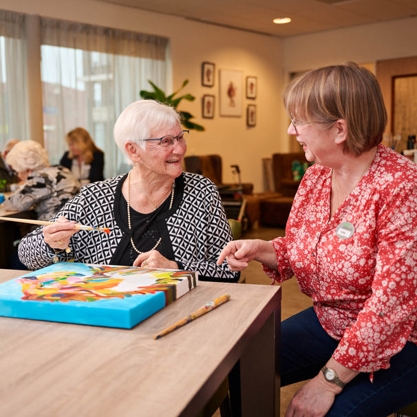 Bewoonster zit met activiteitenbegeleider aan een tafel, voor haar ligt een canvas waarop de bewoonster schildert