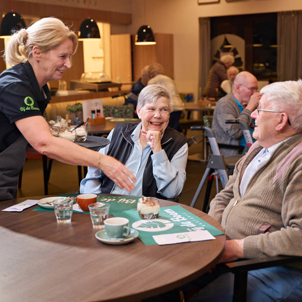 Oudere meneer en mevrouw zitten aan een tafel, lachen naar elkaar terwijl een mevrouw toetjes en koffie op tafel zet