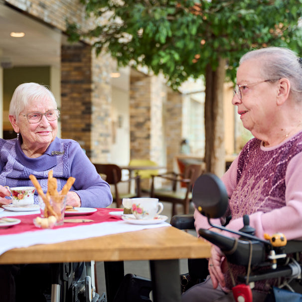 Twee oudere dames zitten aan tafel en drinken koffie, terwijl ze met elkaar kletsen