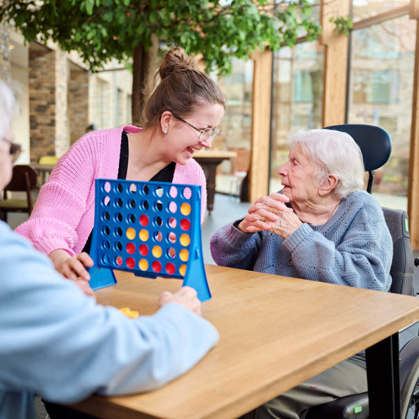 Twee oudere dames zitten tegenover elkaar aan tafel, met daartussenin het spel vier op een rij. Een activiteitenbegeleider staat ernaast en lacht met een van de vrouwen.